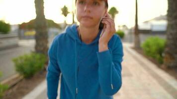 Frau mit Kopfhörer läuft Nieder das Straße entlang das Palme Allee beim Sonnenuntergang. gesund aktiv Lebensstil. schleppend Bewegung video