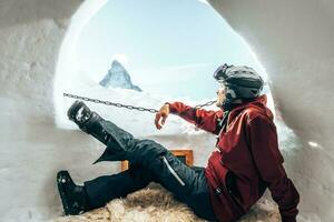 Portrait of smiling tourist snowboarder sitting inside an iglu dorf with a view on the famous snowcapped Matterhorn mountain. Relaxing in Swiss Alps, Zermatt ski resort. photo