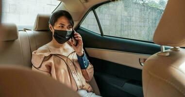 Business woman wearing protective face mask talking by mobile phone while sitting on back seat in the car photo