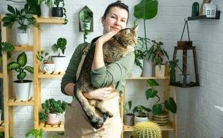 Woman holding pet cat in her arms near her collection of rare species home potted plants on shelf in interior of house. Home plant growing, green house, natural living corner photo