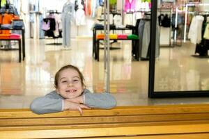 contento pequeño niña en un banco en contra el antecedentes de un de moda ropa Tienda en un centro comercial foto