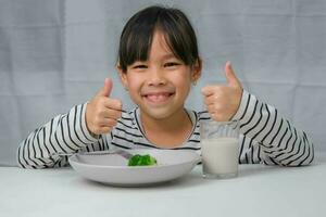 Cute Asian girl drinking a glass of milk in the morning before going to school. Little girl eats healthy vegetables and milk for her meals. Healthy food in childhood. photo