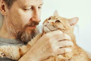 barbado hombre abrazando y acariciando jengibre gato de cerca. selectivo atención en de gato bozal foto