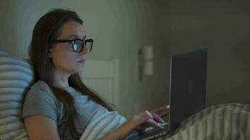 mujer en lentes trabajando en ordenador portátil mientras acostado en cama a noche. móvil adicto o insomnio concepto. video