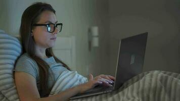 Woman in glasses working on laptop while lying in bed at night. Mobile addict or insomnia concept. video