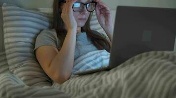 mulher dentro óculos trabalhando em computador portátil enquanto deitado dentro cama às noite. Móvel viciado ou insônia conceito. video