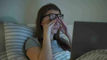Woman in glasses working on laptop while lying in bed at night. Mobile addict or insomnia concept. video