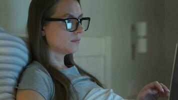 Woman in glasses working on laptop while lying in bed at night. Mobile addict or insomnia concept. video