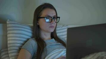 kvinna i glasögon arbetssätt på bärbar dator medan liggande i säng på natt. mobil missbrukare eller sömnlöshet begrepp. video