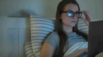 Woman in glasses working on laptop while lying in bed at night. Mobile addict or insomnia concept. video