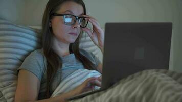 mujer en lentes trabajando en ordenador portátil mientras acostado en cama a noche. móvil adicto o insomnio concepto. video