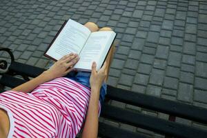 el niña es sentado en un parque banco y leyendo un libro. parte superior ver de un joven niña leyendo un libro. foto