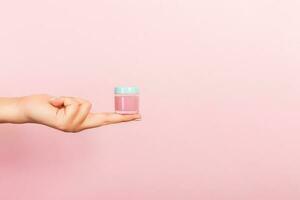 Female hand holding cream bottle of lotion isolated. Girl give jar cosmetic products on pink background photo