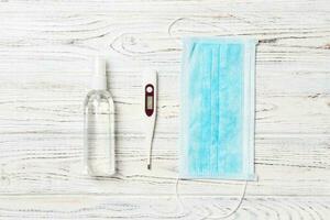 Top view of hygienic surgical mask, digital thermometer and alcohol gel sanitizer on wooden background. Protective equipment concept with copy space photo