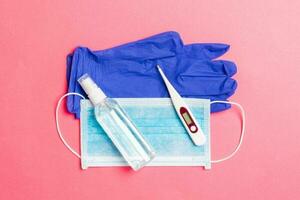 Top view of alcohol hand sanitizer, latex gloves, digital thermometer and medical mask on pink background. Virus protection equipment concept with copy space photo