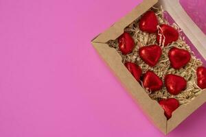 un grupo de en forma de corazon chocolates envuelto en rojo frustrar, acostado en un regalo caja en un rosado antecedentes foto