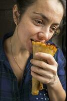 A pretty middle-aged woman in a purple plaid shirt is eating a pizza cone. Fast food. photo