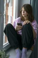 Cute smiling Caucasian teen girl is sitting on the windowsill and looking out the window photo