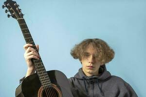 a teenager in a gray hoodie and wavy hair is holding a guitar on a blue background photo