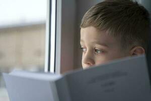 A cute Caucasian boy of 5 years old in a red T-shirt sits on the windowsill by the window with a book photo