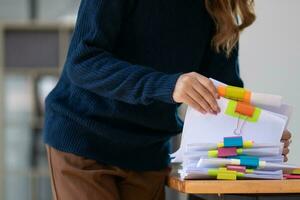 mujer de negocios manos trabajando en pilas de papel documentos a buscar y revisión documentos apilado en mesa antes de enviando ellos a tablero de directores a utilizar correcto documentos en reunión con empresario foto