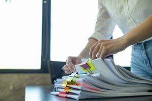 mujer de negocios manos trabajando en pilas de papel documentos a buscar y revisión documentos apilado en mesa antes de enviando ellos a tablero de directores a utilizar correcto documentos en reunión con empresario foto