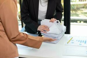 mujer de negocios manos trabajando en pilas de papel documentos a buscar y revisión documentos apilado en mesa antes de enviando ellos a tablero de directores a utilizar correcto documentos en reunión con empresario foto