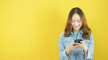 Stylish Asian woman in jeans chatting social media on yellow background. video