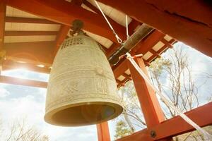 Big brass Buddhist bell and timber knock of Japanese temple in red pavilion on bright blue sky with sun and lens flare background. photo