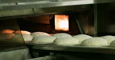 een bakker plaatsen de rauw deeg broodjes binnen een voorverwarmd dek oven gebruik makend van een lader riem - bakken brood in een bakkerij - detailopname schot video