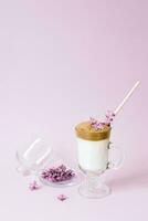 Beautiful drink dalgone coffee with foam and a paper tube in a transparent Cup and lilac flowers on a purple or lilac background. Next to the mug, on a serving platter, is a branch photo