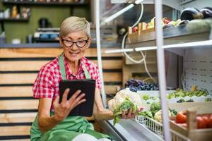 Senior woman works in fruits and vegetables shop. She is holding a tablet device and a cauliflower.. photo