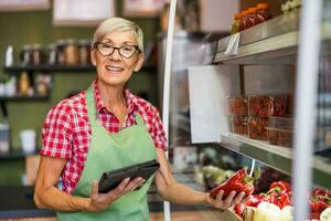 Senior woman works in fruits and vegetables shop. She is holding a tablet device and a red pepper.. photo