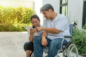 The patient caused by a car accident sitting in a wheelchair and his wife is beside And holding a tablet Let him look to relax while strolling in the garden. Concept of being by side and encouraging. photo