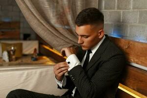 Handsome young man in a stylish suit corrects cufflinks. Guy is resting in a bar. photo