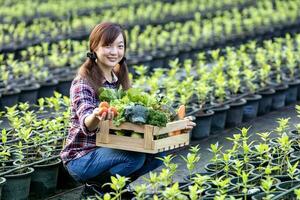 asiático mujer granjero es mostrando el de madera bandeja lleno de recién recoger orgánicos vegetales en su jardín para cosecha temporada y sano dieta alimento. foto