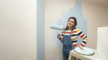 mujer en pie vestido en mono, sonriente a cámara con azul pintar rodillo en mano en el medio de vacío habitación con medio pintado blanco pared antecedentes foto