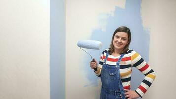 Beautiful woman dressed in overalls, smiling at camera with blue paint roller in her hand against half-painted white wall background photo
