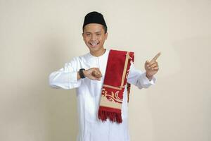 retrato de atractivo asiático musulmán hombre en blanco camisa mirando en muñeca reloj mientras señalando lejos con su dedo. aislado imagen en gris antecedentes foto