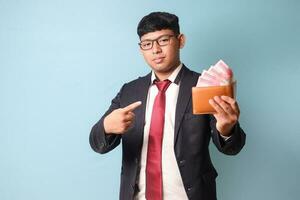 Portrait of young Asian business man in casual suit holding and pointing at leather wallet with thousand rupiahs. Isolated image on blue background photo