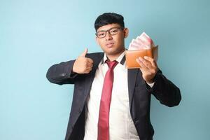 Portrait of young Asian business man in casual making thumb up while holding leather wallet with thousand rupiahs. Isolated image on blue background photo