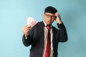 Portrait of young Asian business man in casual suit scartching head looking confused while holding thousand rupiahs. Isolated image on blue background photo