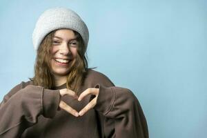 un linda europeo Adolescente niña en de moda de punto sombrero doblada su corazón fuera de su dedos y sonrisas foto
