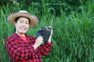 asiático mujer agricultor, sostiene soy, fm transistor radio receptor en hombro. concepto contento trabajando a lo largo con música, noticias, información ,anuncio desde radio. país estilo de vida. término análogo tecnología. foto