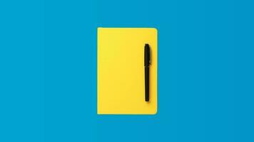 Photo of closed yellow notebook and ball-point pen over blue background. Minimalist image of closed diary and black pen as back to school concept.