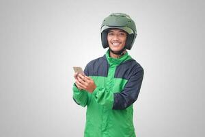 retrato de asiático en línea Taxi conductor vistiendo verde chaqueta y casco participación un móvil teléfono y sonriente. publicidad concepto. aislado imagen en blanco antecedentes foto