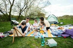 contento joven familia con cuatro niños teniendo divertido y disfrutando al aire libre en picnic cobija pintura a jardín primavera parque, relajación. foto