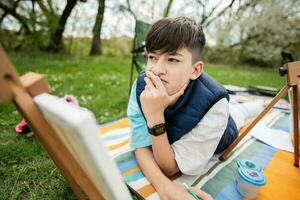 Teenager boy enjoying outdoor on picnic blanket and painting on canvas at garden spring park, relaxation. photo