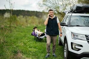 Man in sleeveless vest speak on phone against suv car with roof rack box, family on picnic. photo