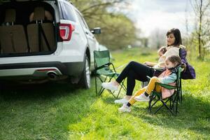 Mother with two daughters sit on chair against car open trunk on picnic. photo